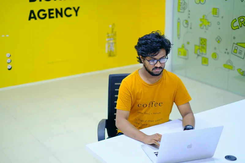 Man working on a laptop at a desk in front of a yellow wall reading “Agency”