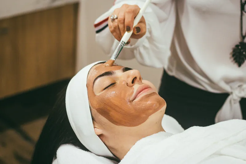 Beautician applying a clay face mask with a brush to a relaxed client