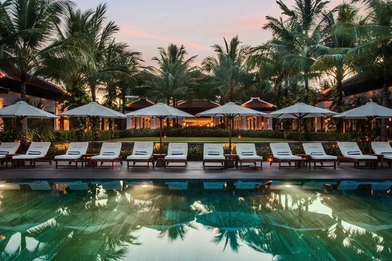 Resort swimming pool at dusk, lined with sun loungers, white parasols and palm trees